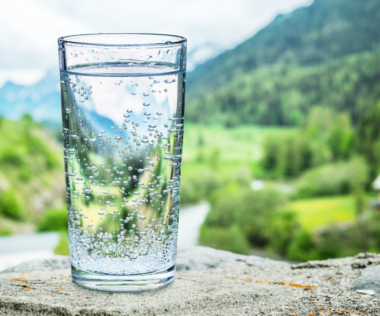 Glass of water on the stone Blurred snow mountains tops and green forests at the background, as a symbol of freshness and purity.